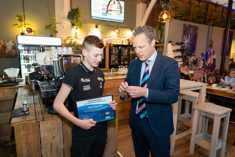 Een wethouder staat in een café naast een jongen. De wethouder reikt een jeugdlintje uit