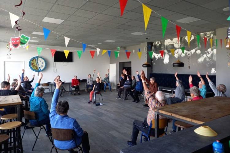 Een groep ouderen zit in op stoelen in een kring en doet mee aan een beweegactiviteit