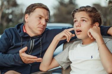 Een man zit buiten naast een puber met zijn hand open in de richting van de puber, terwijl hij naar de puber kijkt. De puber kijkt omhoog en houdt zijn vingers in zijn oren