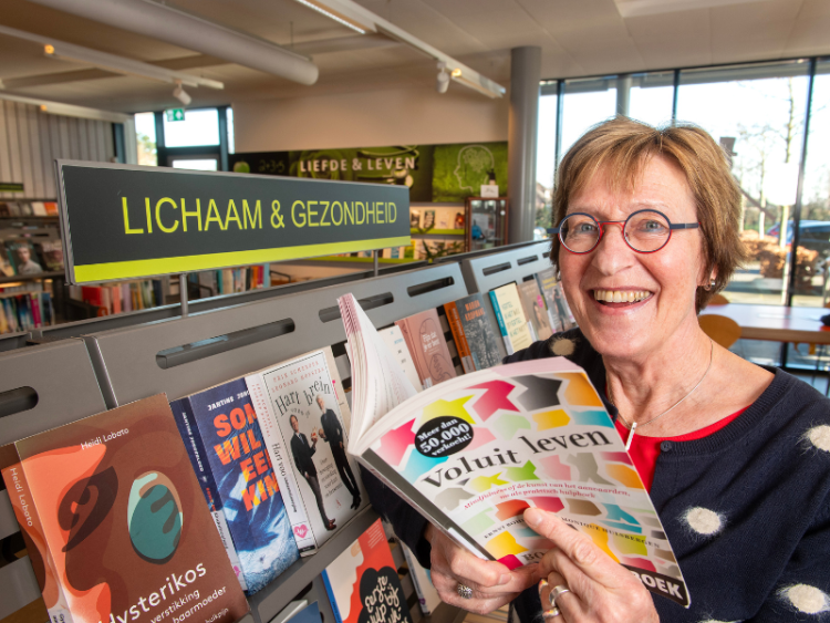 . Els Duijn, leefstijladviseur van de GGD Gelderland-Zuid, staat voor een boekenkast en houdt een boek vast met als titel 'Voluit leven'