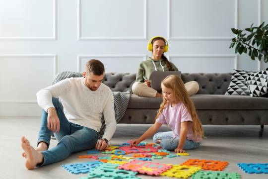 Decoratieve afbeelding: Een foto van een gezin. Moeder zit op de bank en vader legt een vloerpuzzel met zijn dochter.