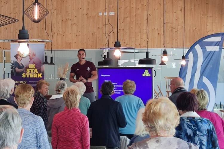 Een man met een microfoontje voor zijn mond staat in een zaal op een verhoging terwijl een groep ouderen naar hem kijkt en een beweegactiviteit doet