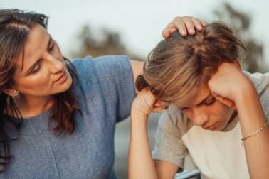 Een tiener zit met het hoofd omlaag en leunt op zijn armen, terwijl een vrouw hem met haar hand over zijn hoofd streelt