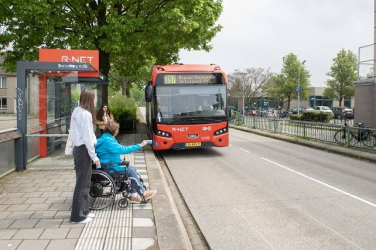 OV-coach Layla Giraud oefent een route in het openbaar vervoer met een deelnemer die in een rolstoel zit. Samen wachten ze op een busplatform op een bus van R-Net die er net aan komt rijden.
