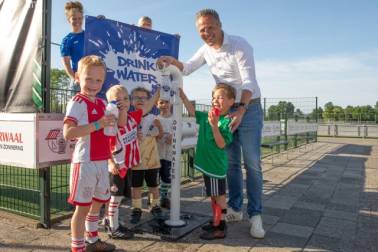 5 kinderen staan samen met wethouder Rutger van Stappershoef bij een watertappunt bij een sportveld. 2 kinderen houden een bidon vast.