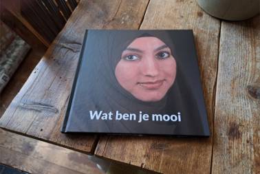Een boek ligt op een houten tafel. Het boek heeft als titel 'Wat ben je mooi' en als omslagfoto een vrouw met hoofddoek die in de camera kijkt.