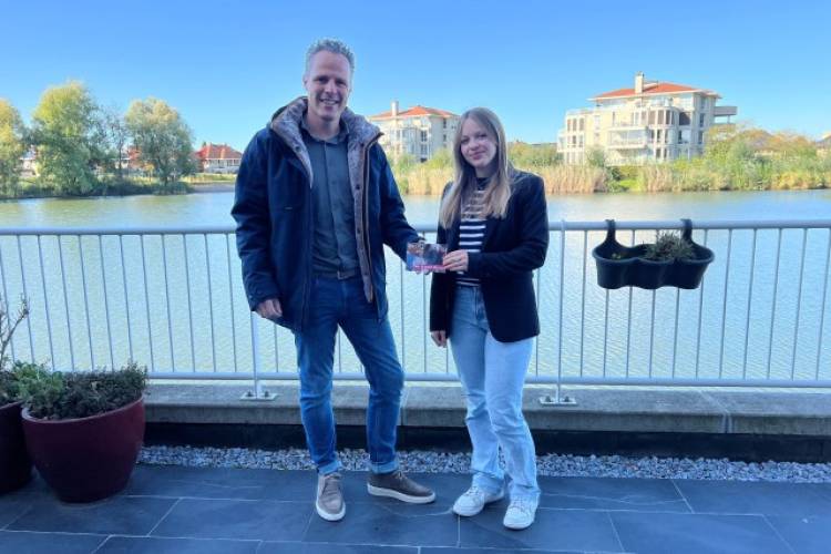 Melica Vermeulen uit Geldermalsen krijgt een verjaardagskaart Yes 18 van wethouder Rutger van Stappershoef, terwijl beide personen voor een hek staan met water daarachter