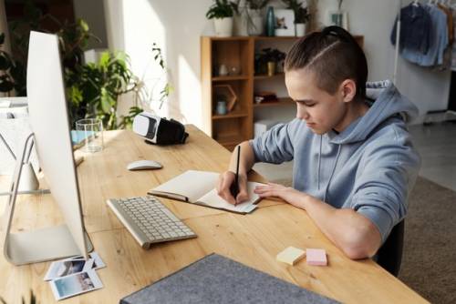 Een tiener met capuchontrui noteert iets in een opschrijfboek terwijl hij achter een computer zit