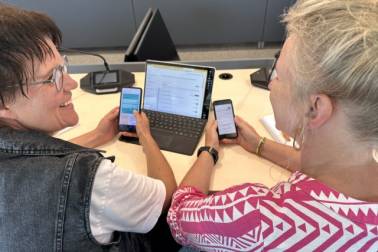 2 budgetcoaches van de gemeente West Betuwe zitten elk met een smartphone in hun hand aan een bureau waar een laptop op staat.