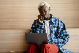 Een jongvolwassene zit met een laptop op schoot en is tegelijkertijd aan het bellen