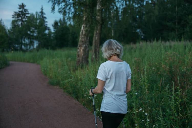 Een vrouw loopt met een wandelstok over een pad langs hoog gras, op de rug gezien