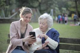 Een jongere vrouw laat aan een oudere vrouw iets zien op haar telefoon, terwijl ze op een bankje zitten
