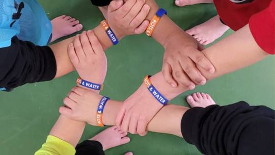 Close up van een groep kinderen die handen vasthouden. Ze hebben Rots en Water-polsbandjes om.