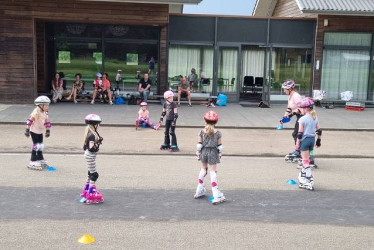 Kinderen met helmen op en beschermers om krijgen buiten skateles, terwijl een aantal volwassenen toekijken.