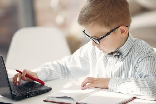 Een kind van rond de 10 jaar oud kijkt door zijn bril naar een laptopscherm, met een opschrijfboek voor zich op tafel