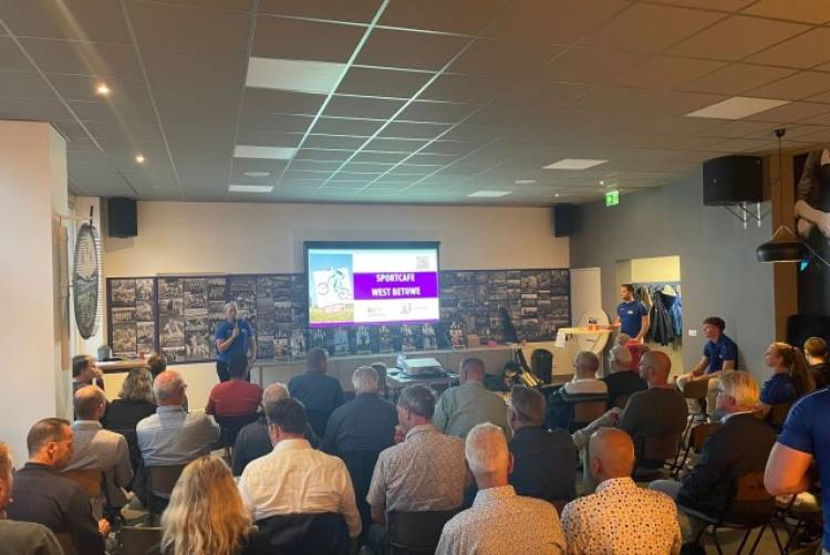 2 mensen met een blauw t-shirt aan staan voorin een zaal bij een groot scherm met daarop de tekst 'Sportcafé West Betuwe'. In de zaal zitten tientallen mensen op klapstoelen.