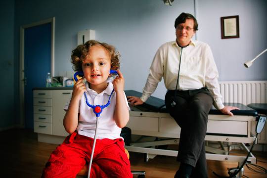 Een jongetje met een stethoscoop in zijn oren en de huisarts in de achtergrond