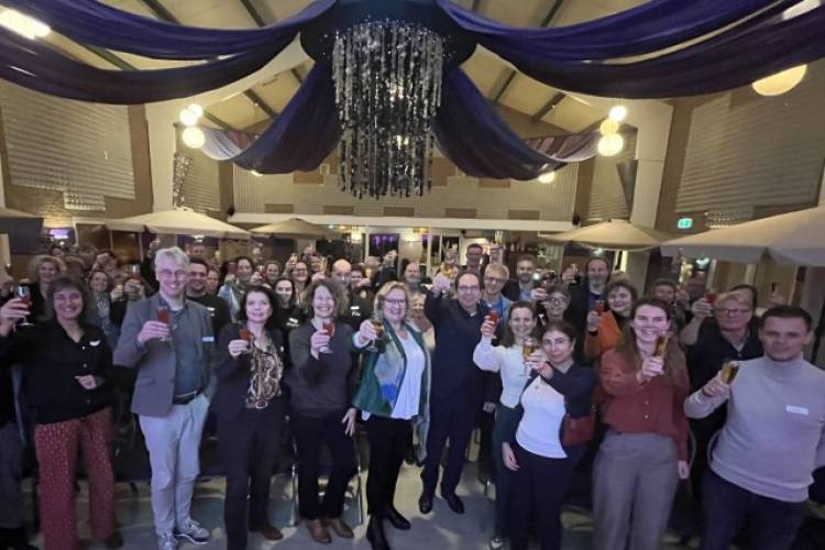Wethouders Wil Kosterman en Jan de Geus staan met een grote groep mensen in een zaal en houden een gevuld glas omhoog