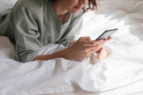 Een jonge vrouw waarvan het gezicht niet helemaal te zien is, ligt op haar buik op een bed terwijl ze haar mobiele telefoon bedient.